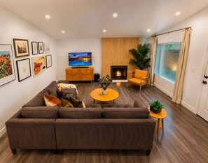 A living room with a sectional sofa, a flat-screen TV, a fireplace, and a large window with blinds. - 25% Off Oct 22-25 Perfect 4 Families Pool, Jacuzzi (Palm Springs)