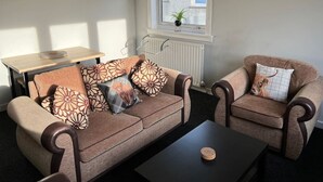 A living room with a sofa, armchair, and coffee table. There is a window with blinds, a radiator, and a small plant on the windowsill. - The Heilan` Coo Apartment (Argyll and Bute Council)