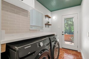 A laundry area with a Samsung washer and dryer, a white countertop, a window with blinds, and a door leading to an outdoor area. - SoCo BoHo Bungalow | Main House (Austin)