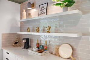 A kitchen with white marble countertops, two white shelves with decorative items, a coffee machine, and a tray with coffee cups and condiments. - SoCo BoHo Bungalow | Main House (Austin)