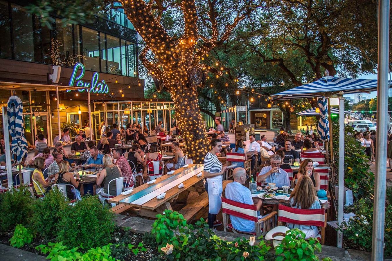 An outdoor dining area with wooden tables and chairs, surrounded by greenery and string lights.