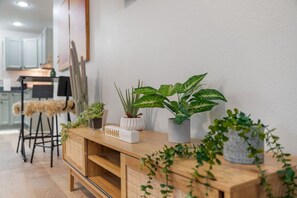 A wooden console table with potted plants and a white object, likely a speaker, in a modern kitchen setting with white cabinets and a bar stool.