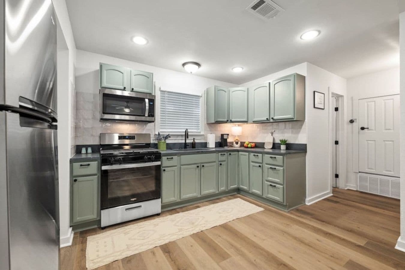 A modern kitchen with stainless steel appliances, light green cabinets, and a wooden floor.