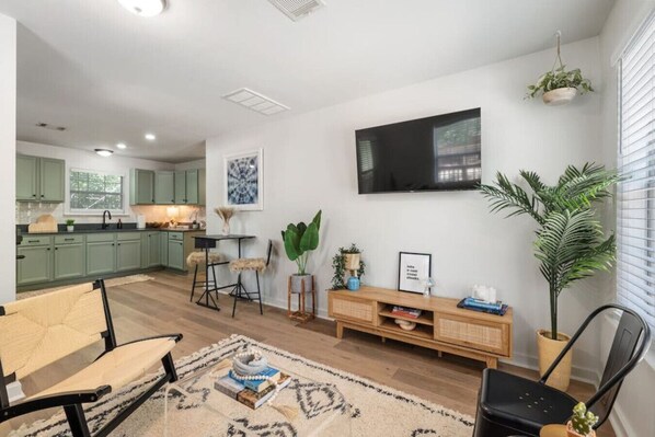 A modern living room with a sofa, a coffee table, and a TV mounted on the wall. There is a kitchen area with green cabinets and a dining table with chairs.