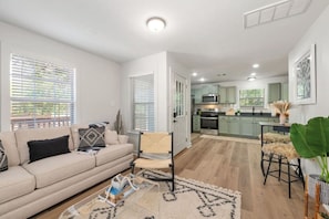 A modern living room with a sofa, a coffee table, and a chair. There is a kitchen area with a microwave, stove, and bar stools.