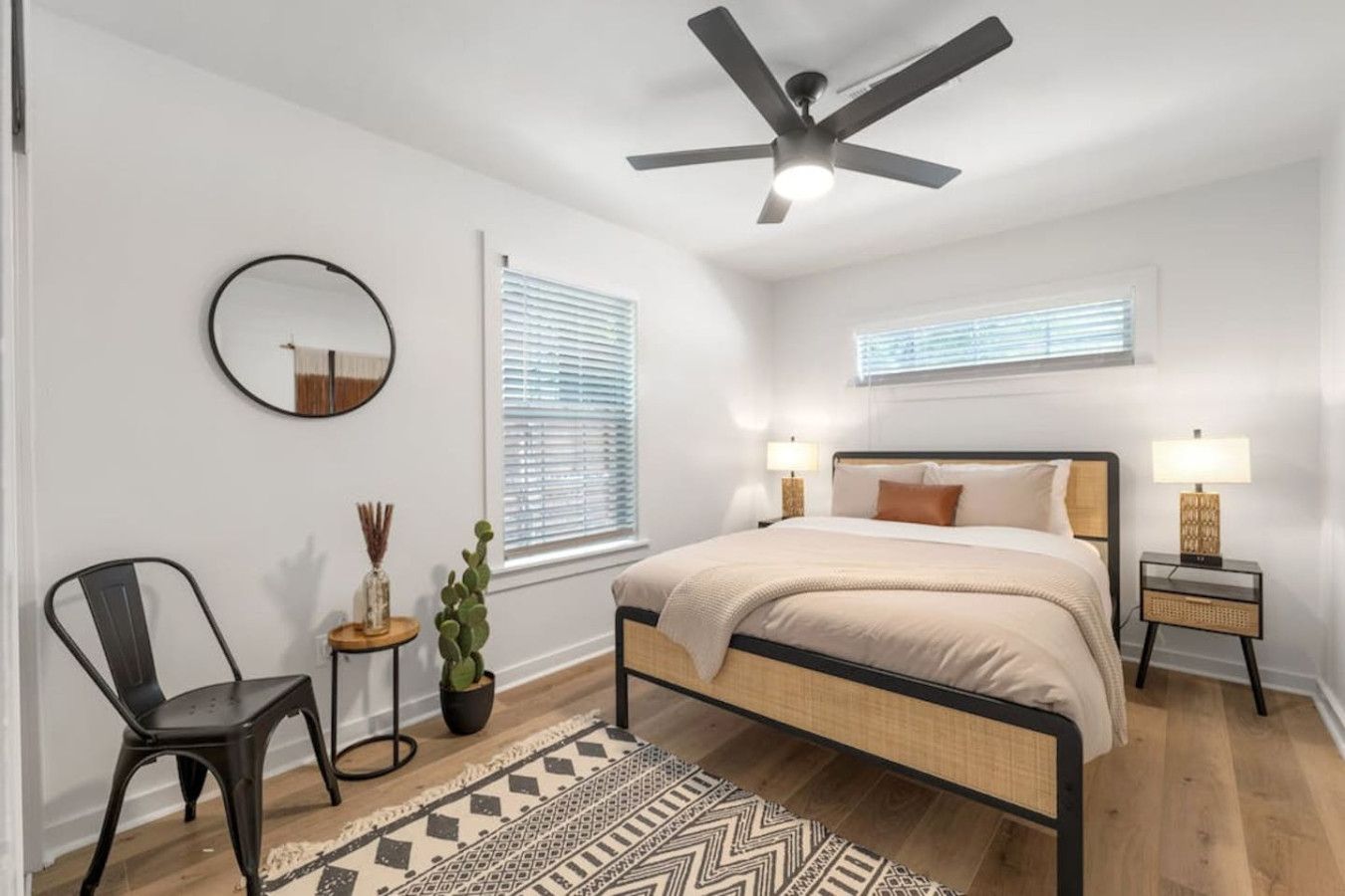 A bedroom with a bed, two bedside tables with lamps, a ceiling fan, a round mirror, a potted plant, a chair, and a patterned rug.