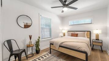 A bedroom with a bed, two bedside tables with lamps, a ceiling fan, a round mirror, a potted plant, a chair, and a patterned rug.