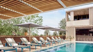 Un espace aménagé au bord de la piscine, avec des chaises longues et des parasols, une piscine à l’eau claire et, en arrière-plan, une structure de bâtiment moderne.