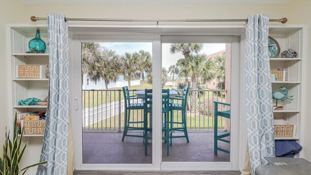 Une pièce dotée d’une porte-fenêtre coulissante donnant sur un balcon où se trouvent deux chaises vertes, et d’où l’on peut admirer des palmiers et une clôture.