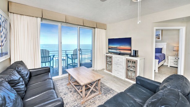 A living room with a sofa, a coffee table, and a TV. There is a balcony with a view of the ocean.