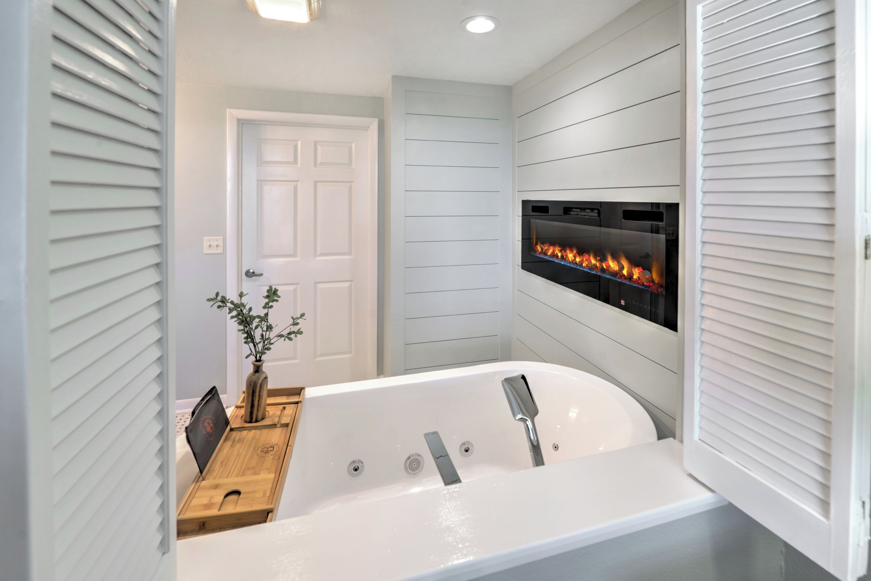 A modern bathroom with a freestanding bathtub, a wall-mounted fireplace, and a wooden side table with a vase.