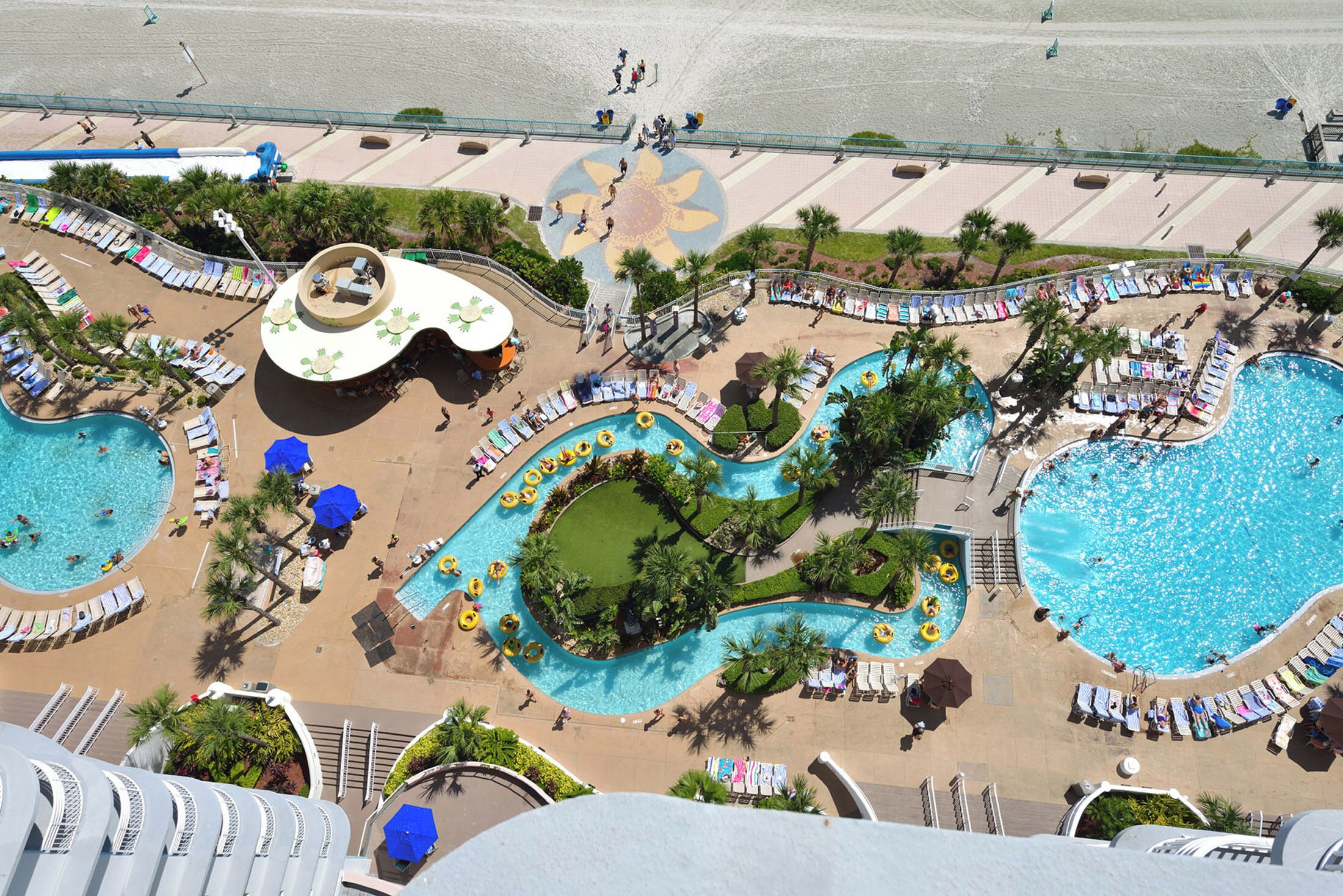 An aerial view of a resort with multiple swimming pools, sun loungers, and a central circular pool surrounded by palm trees.