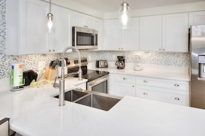 A modern kitchen with white cabinetry, stainless steel appliances, a double sink, and a tiled backsplash. - Oceanfront Escape | King Bed + 2 Balconies (Daytona Beach)