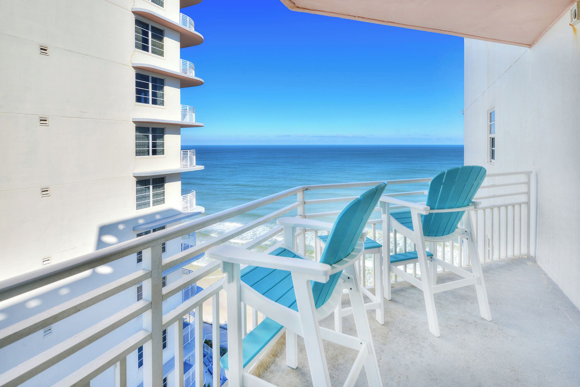 A balcony with two chairs and a table, overlooking a clear blue ocean and a building with multiple balconies.