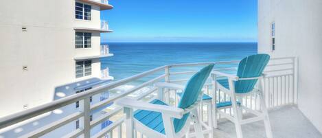 A balcony with two chairs and a table, overlooking a clear blue ocean and a building with multiple balconies.
