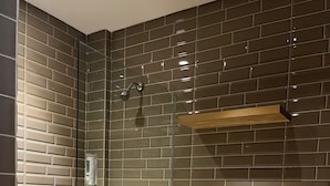 A modern bathroom with a glass-enclosed shower, a white toilet, and a tiled wall with a brick pattern. - Lugano Arte Homestay (Jakarta)