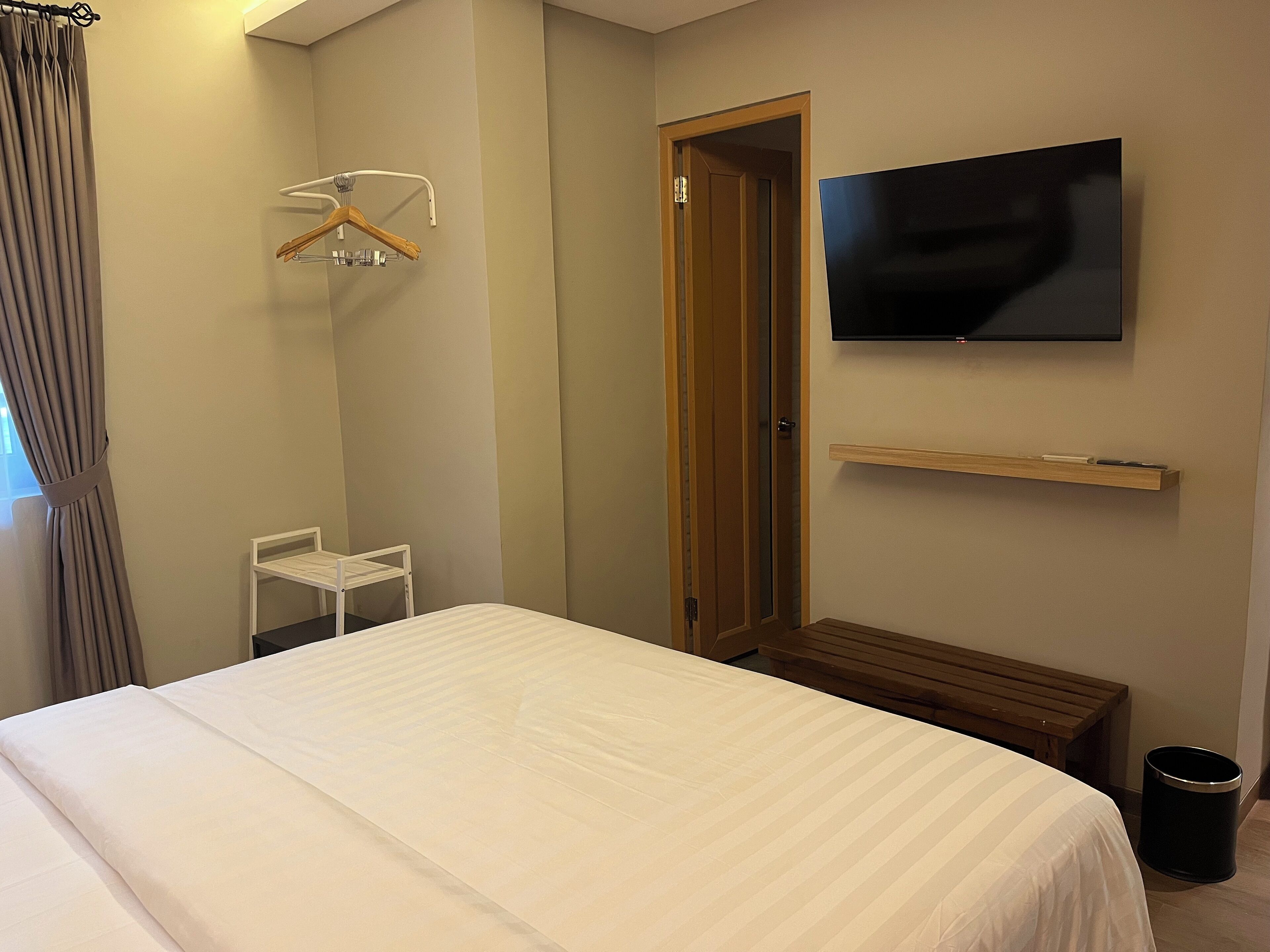 Quarto de hotel com cama, televisão fixada na parede, um banquinho de madeira, uma mesinha, uma lixeira e uma porta.