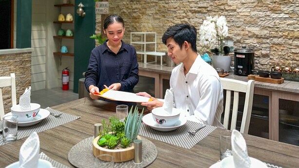 A dining area with a stone wall, a wooden table, and a tree providing shade. - Lugano Arte Homestay (Jakarta)