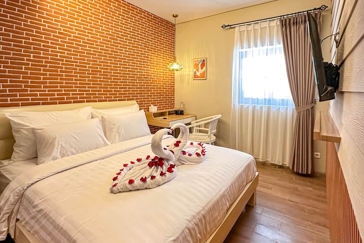A hotel room with a brick wall, a bed with white bedding, a wooden nightstand, a chair, and a window with curtains.