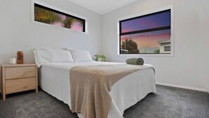 A bedroom with a bed, a nightstand, a window with a view, and a framed picture on the wall. - Another on Leo! (Waihi Beach)