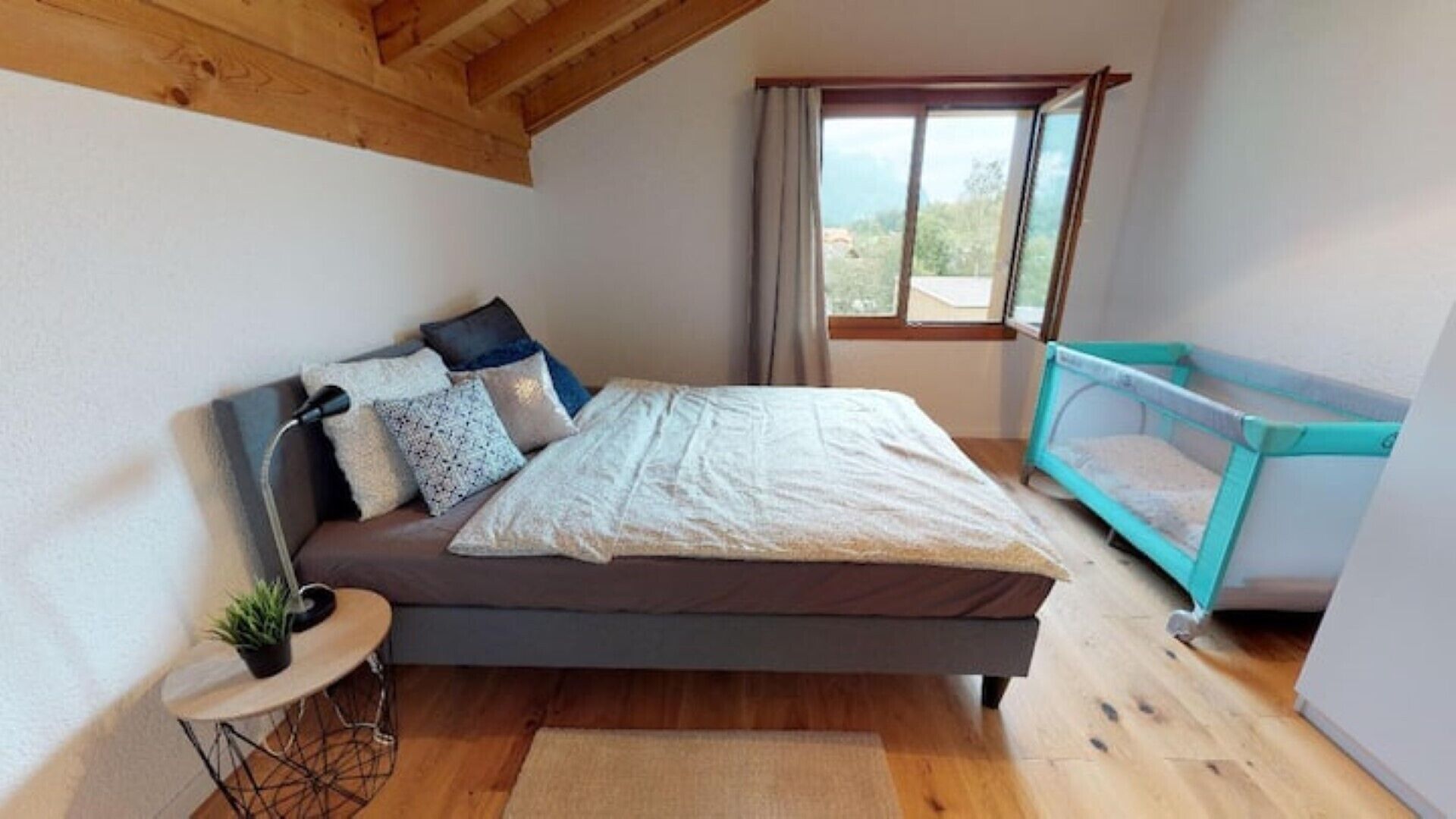 A bedroom with a bed, pillows, a small table with a plant, a window with curtains, and a crib.