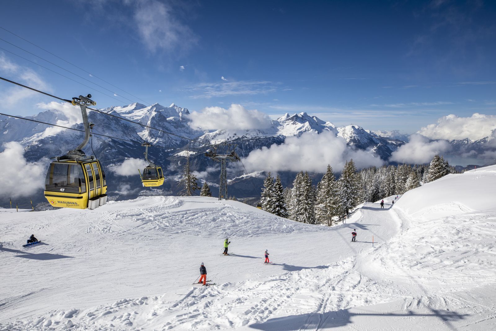 Un télésiège se déplace le long d’une pente de montagne enneigée, avec des skieurs et des snowboardeurs en contrebas.