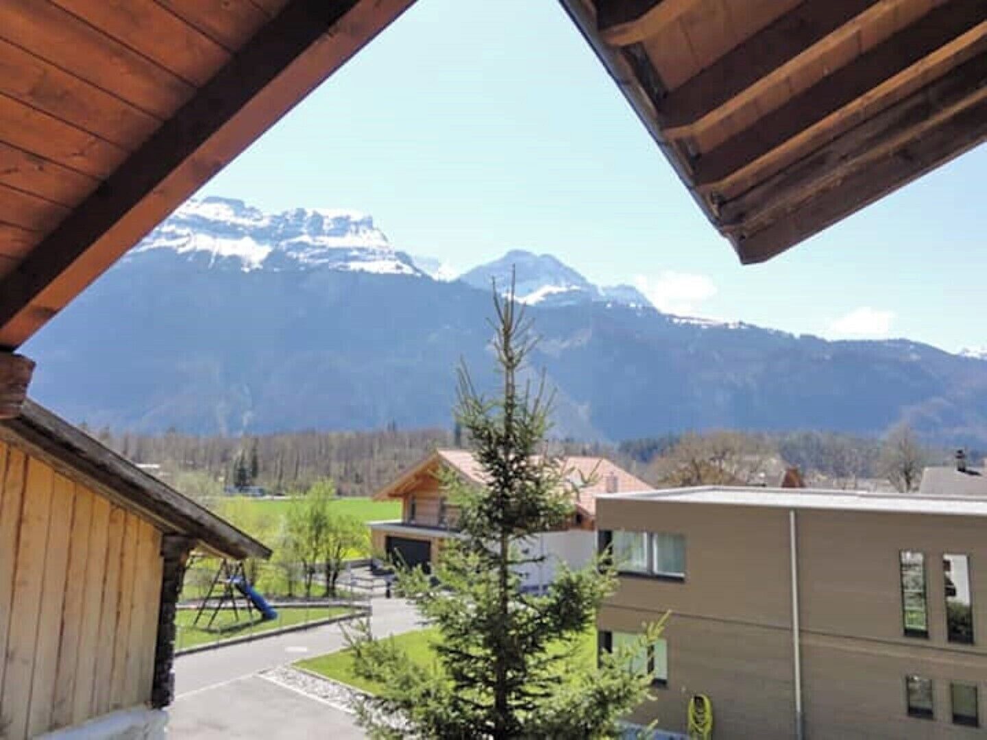 Blick vom Holzbalkon auf ein Gebirge mit einem Spielplatz und modernen Gebäuden im Vordergrund.