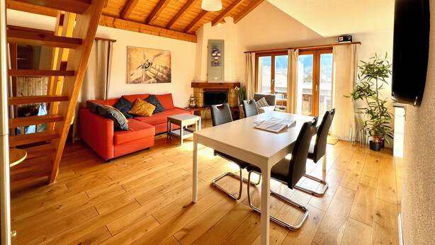 A spacious living room with a red sofa, a dining table with chairs, a staircase, and a large window with a view. - MAISONETTE 4.5 rooms, near Ballenberg, ski areas, playground, BeO ApartmentsAG (Hofstetten bei Brienz)