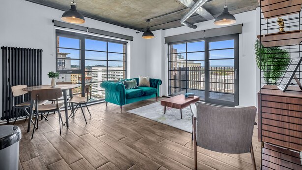 A modern living room with a teal sofa, wooden flooring, and large windows offering a city view. - Wembley Park Apartments (Wembley)