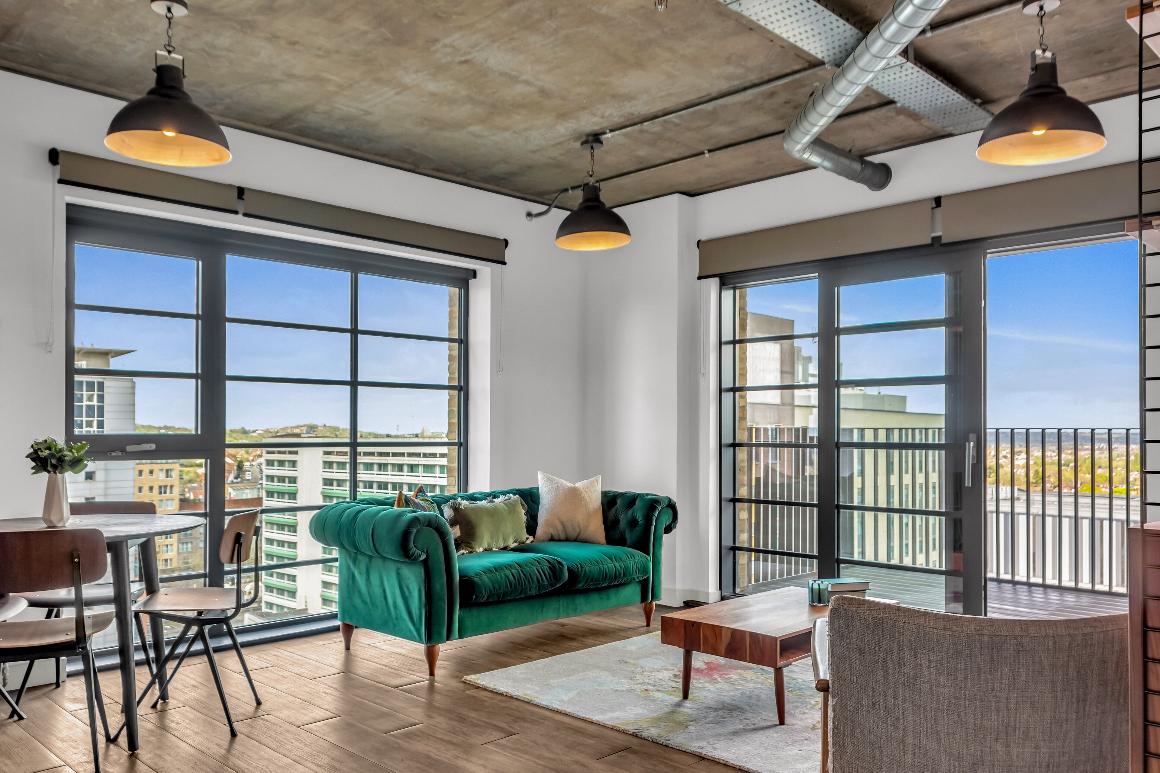 A modern living room with a green sofa, wooden flooring, and large windows offering city views.