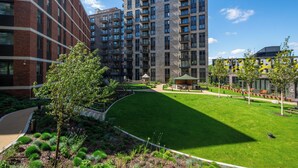 A modern apartment complex with multiple high-rise buildings, a well-maintained courtyard with green lawns, and a variety of trees and shrubs. - Wembley Park Apartments (Wembley)