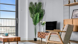 A modern living room with a wooden coffee table, a beige armchair, a large potted plant, a flat-screen TV, and a geometric-patterned cabinet. - Wembley Park Apartments (Wembley)