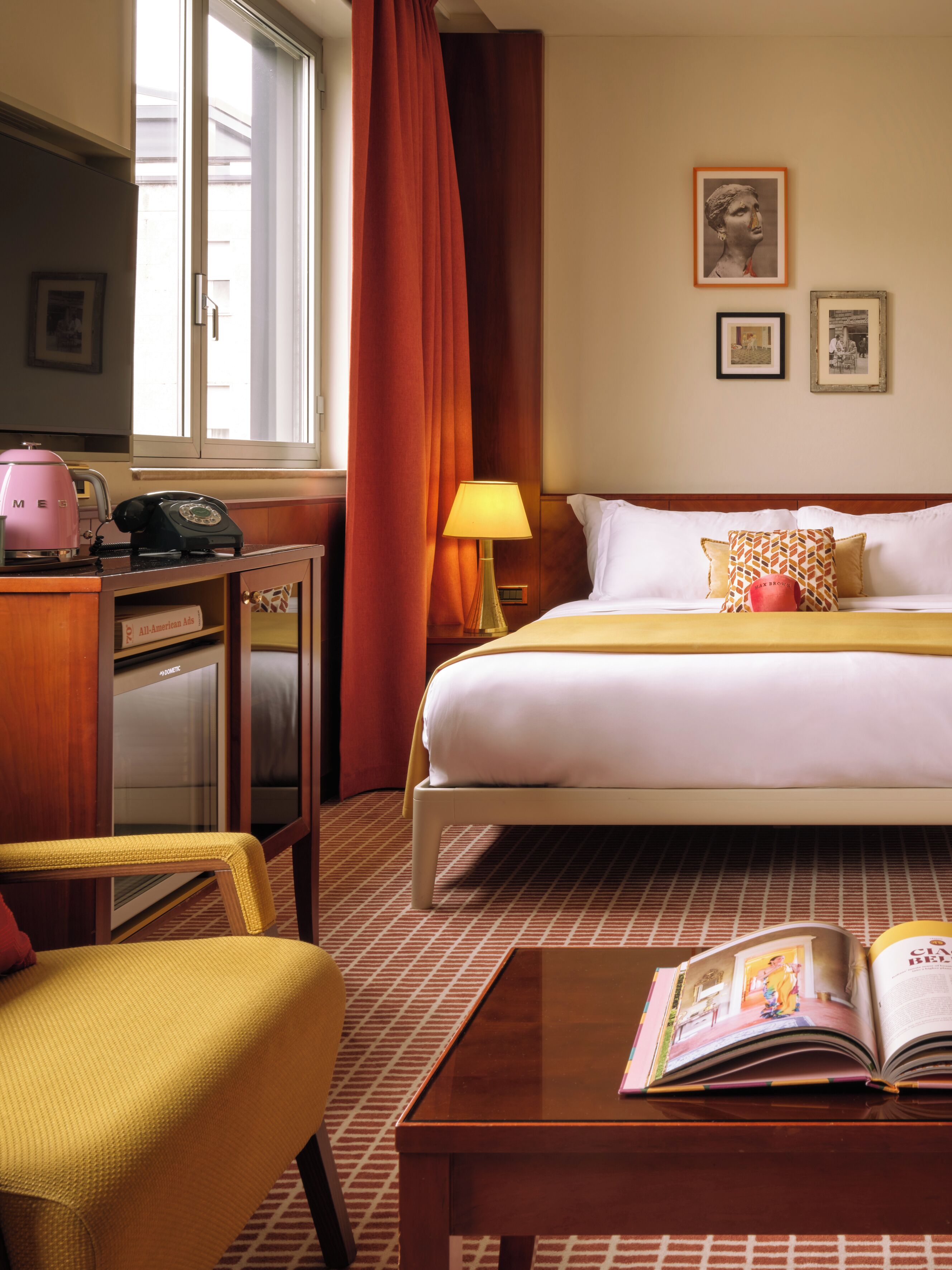 A hotel room with a bed, a desk with a telephone, a chair, a lamp, and a book on a table.