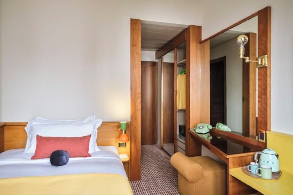 A hotel room with a bed, a bedside table with a lamp, a chair, a desk with a telephone and a kettle, and a mirror.