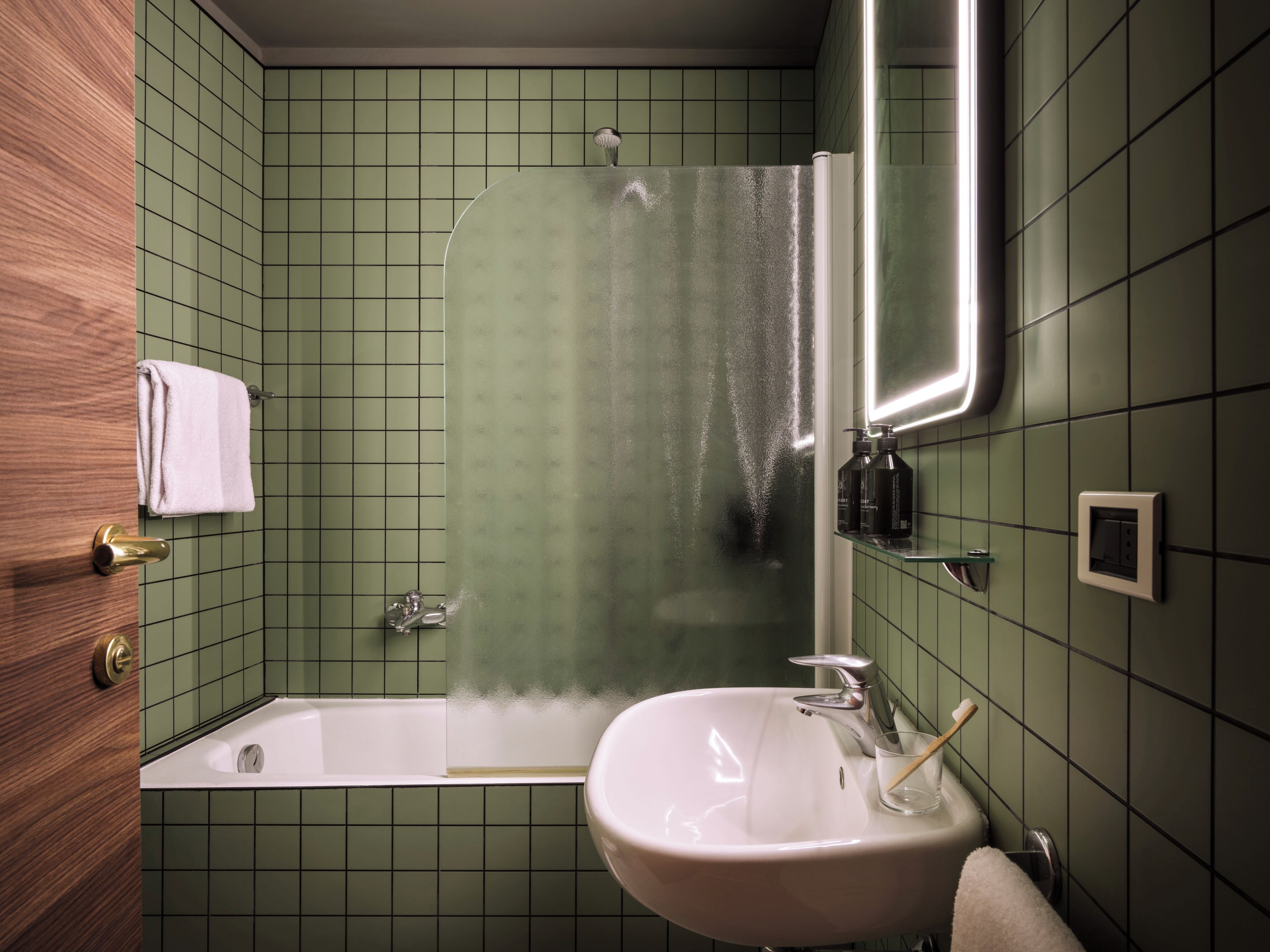 A bathroom with green tiled walls, a white sink, a glass shower enclosure, and a wooden door.