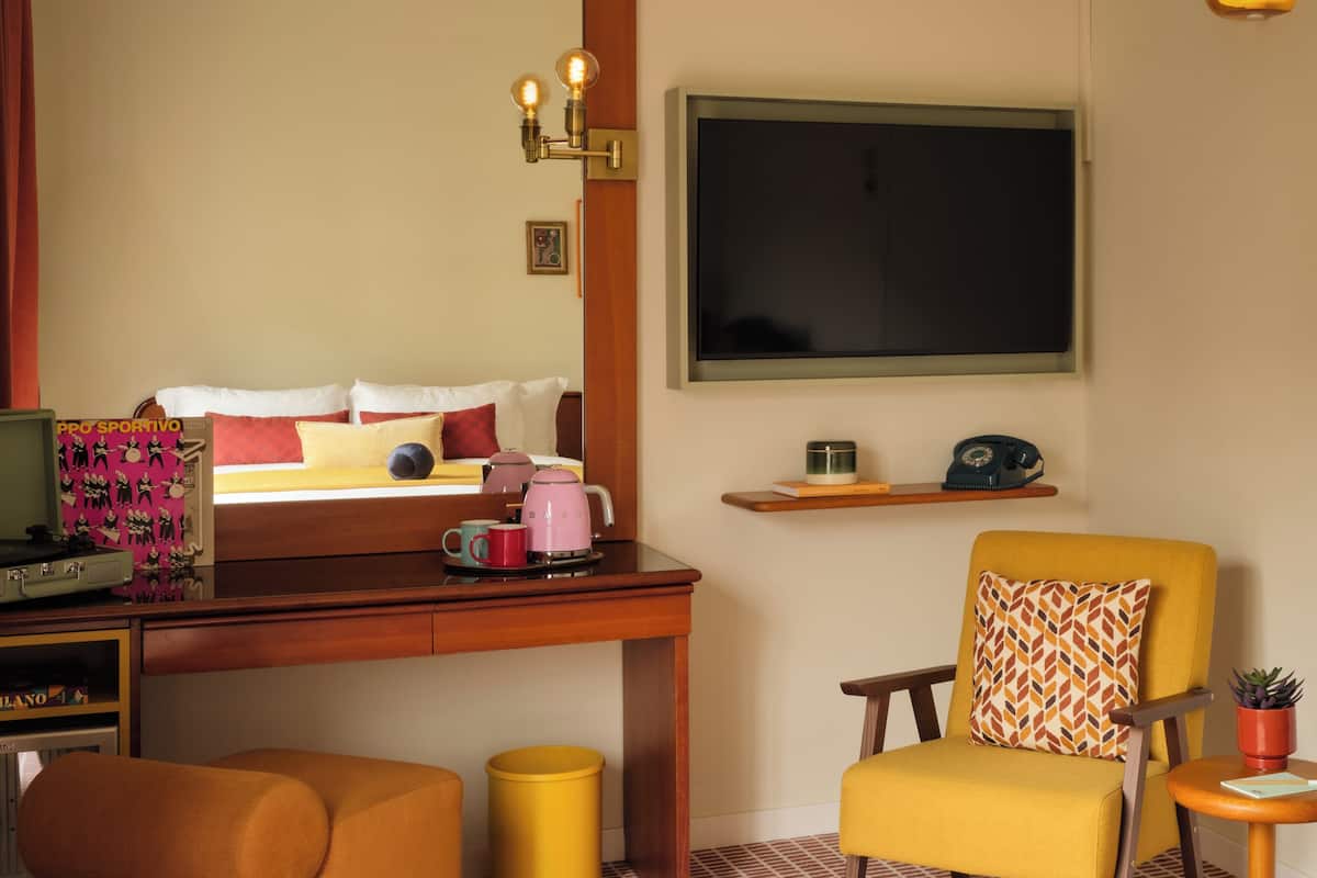A hotel room with a bed, a desk with a computer, a chair, a small table with a plant, a trash can, and a mirror.