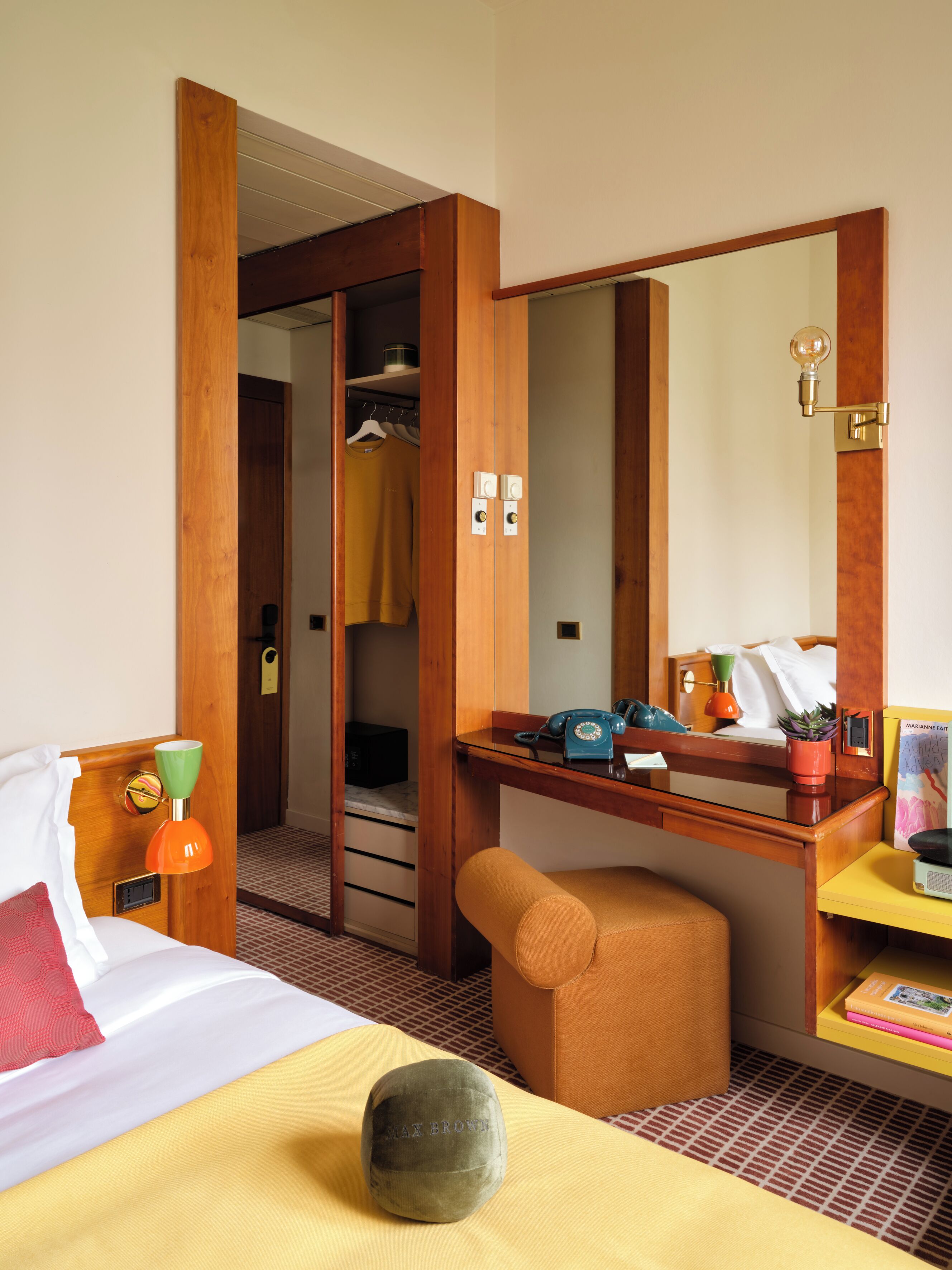 A hotel room with a bed, a desk with a telephone, a mirror, and a small stool.