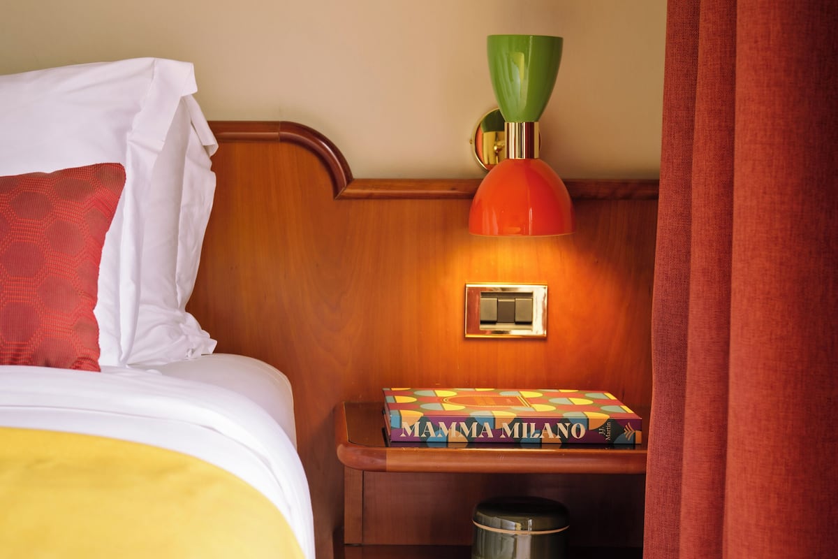 A hotel room with a bed, a nightstand with a lamp, and a book titled "Mamma Milano".