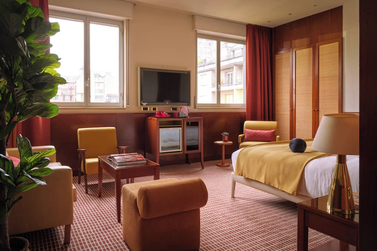 A hotel room with a bed, a TV, a desk, a chair, a sofa, and a potted plant.