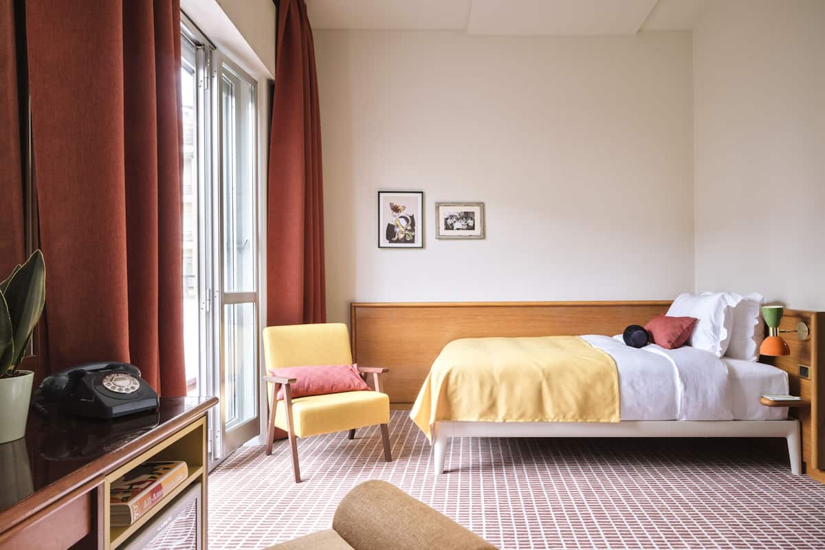 A hotel room with a bed, a chair, a desk with a telephone, and two framed pictures on the wall.
