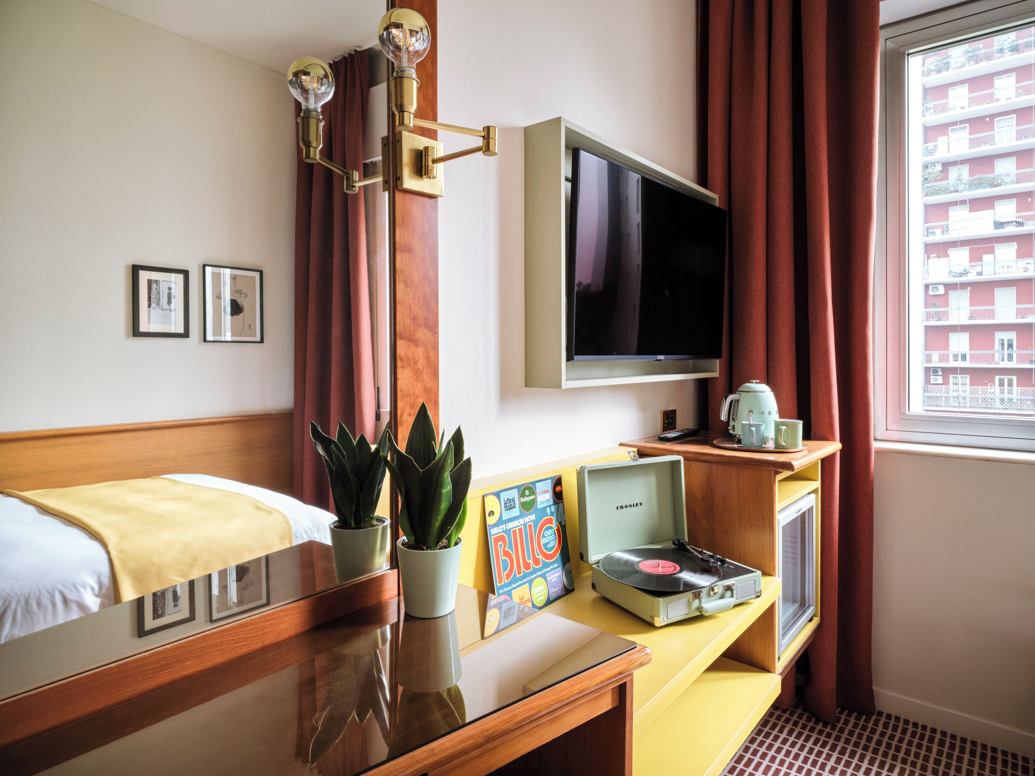 Hotellihuone, jossa on sänky, taulutelevisio, kahvinkeitin, levysoitin ja ikkuna, jossa on verhot.