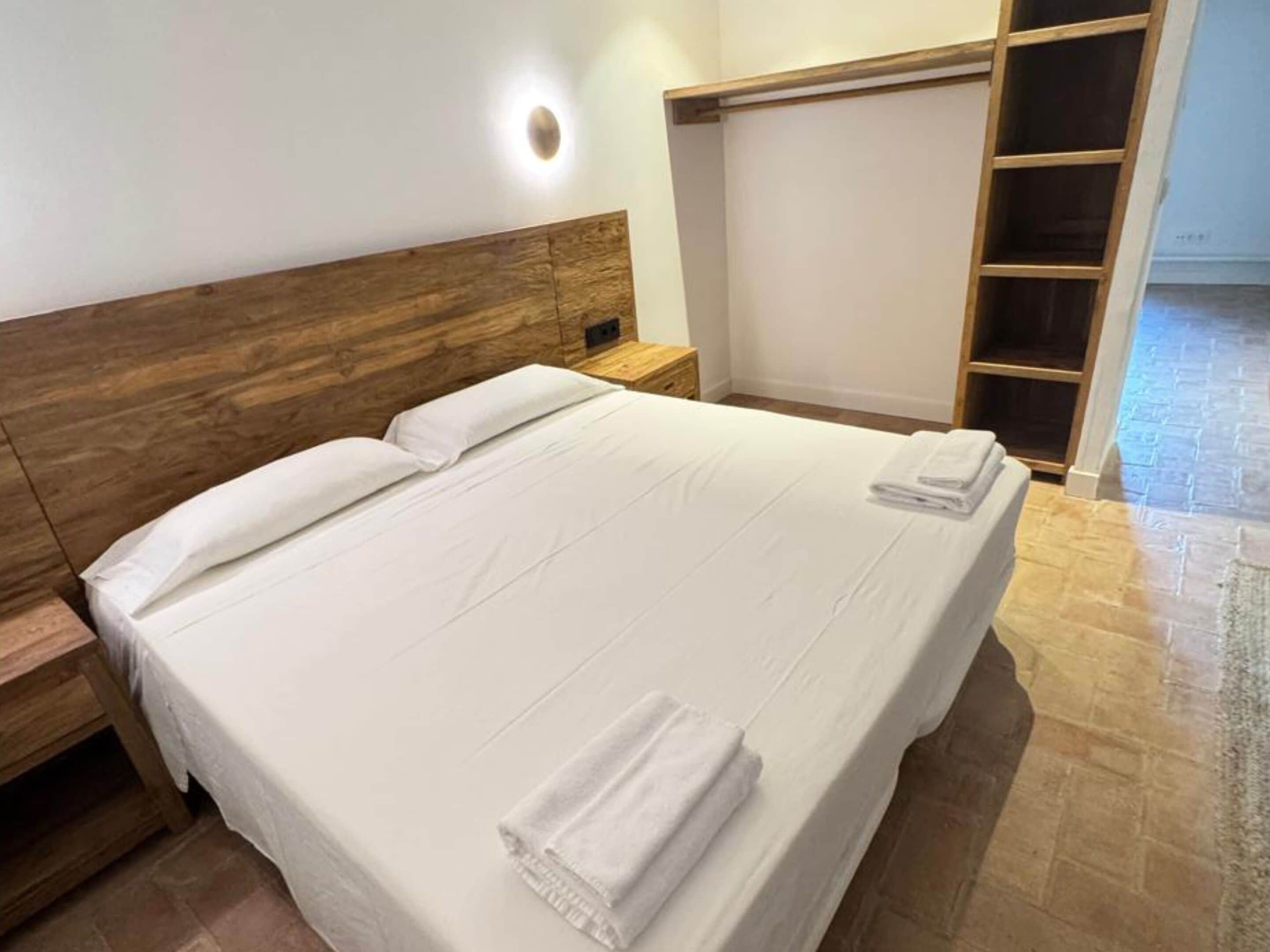Uma cama bem arrumada com lençóis brancos e duas toalhas dobradas, cabeceira de madeira, mesa de cabeceira e guarda-roupa embutido com espelho.