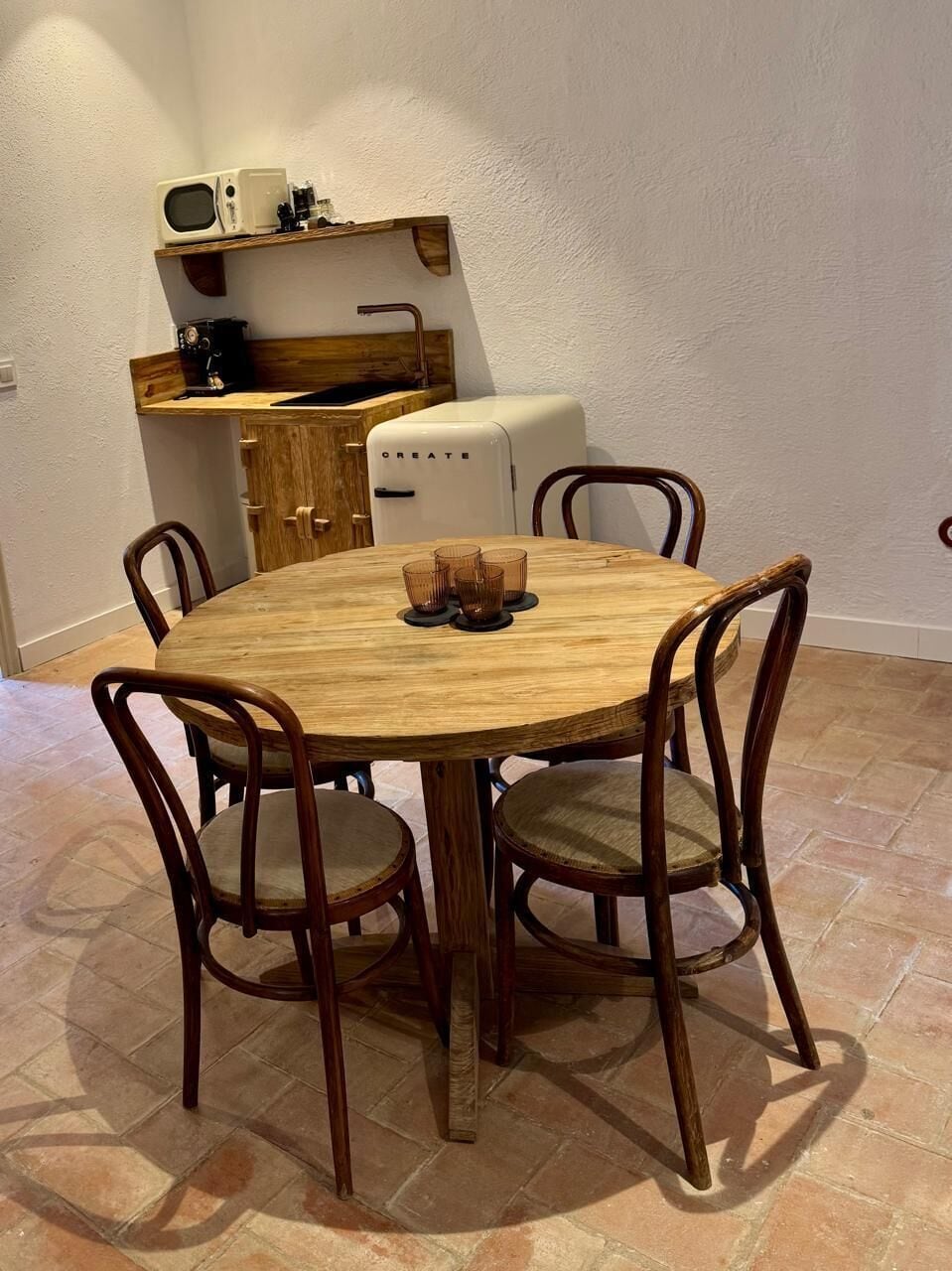 A round wooden dining table with four chairs, a small kitchen area with a microwave and a refrigerator, and a tiled floor.