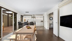 Een moderne keuken met witte kastjes, een eethoek met een houten tafel en stoelen, en een grote televisie aan de muur.