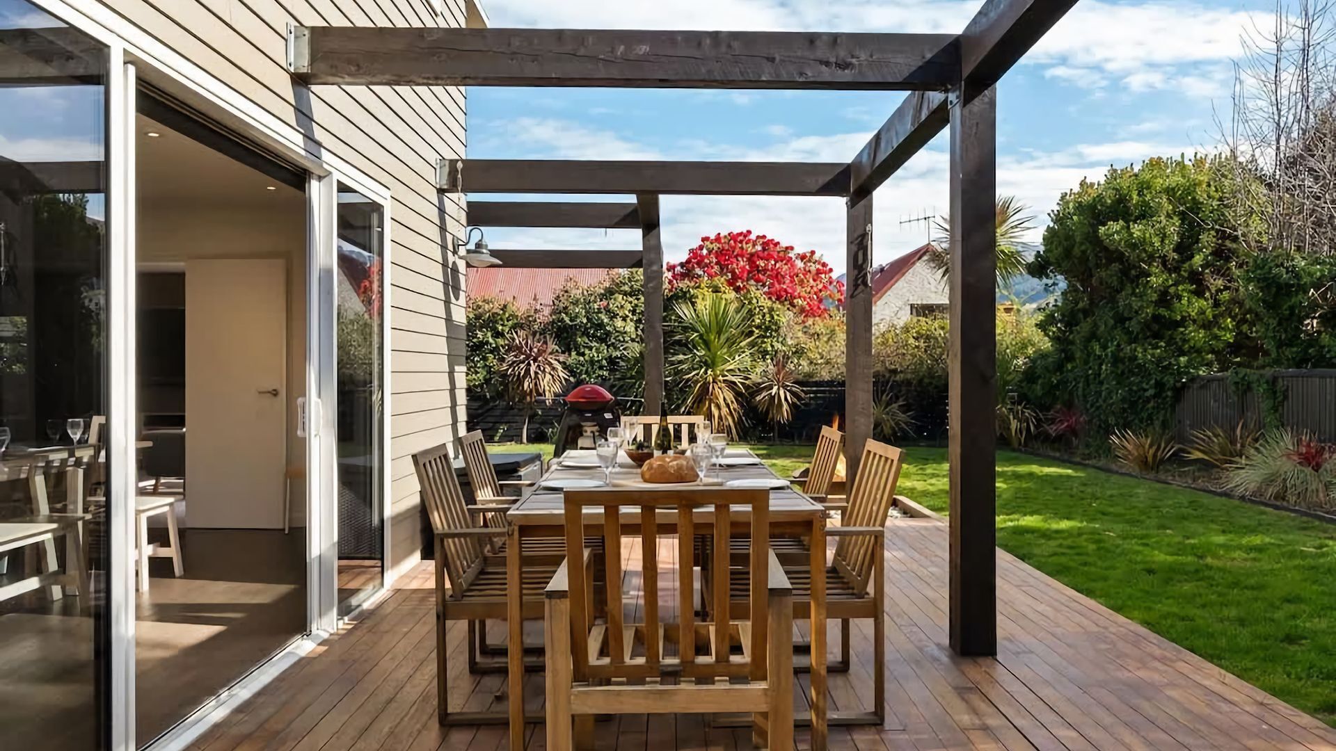 Une terrasse extérieure avec une table et des chaises en bois, une structure en forme de pergola et une vue sur un jardin où poussent différentes plantes, ainsi qu’un arbuste à fleurs rouges.