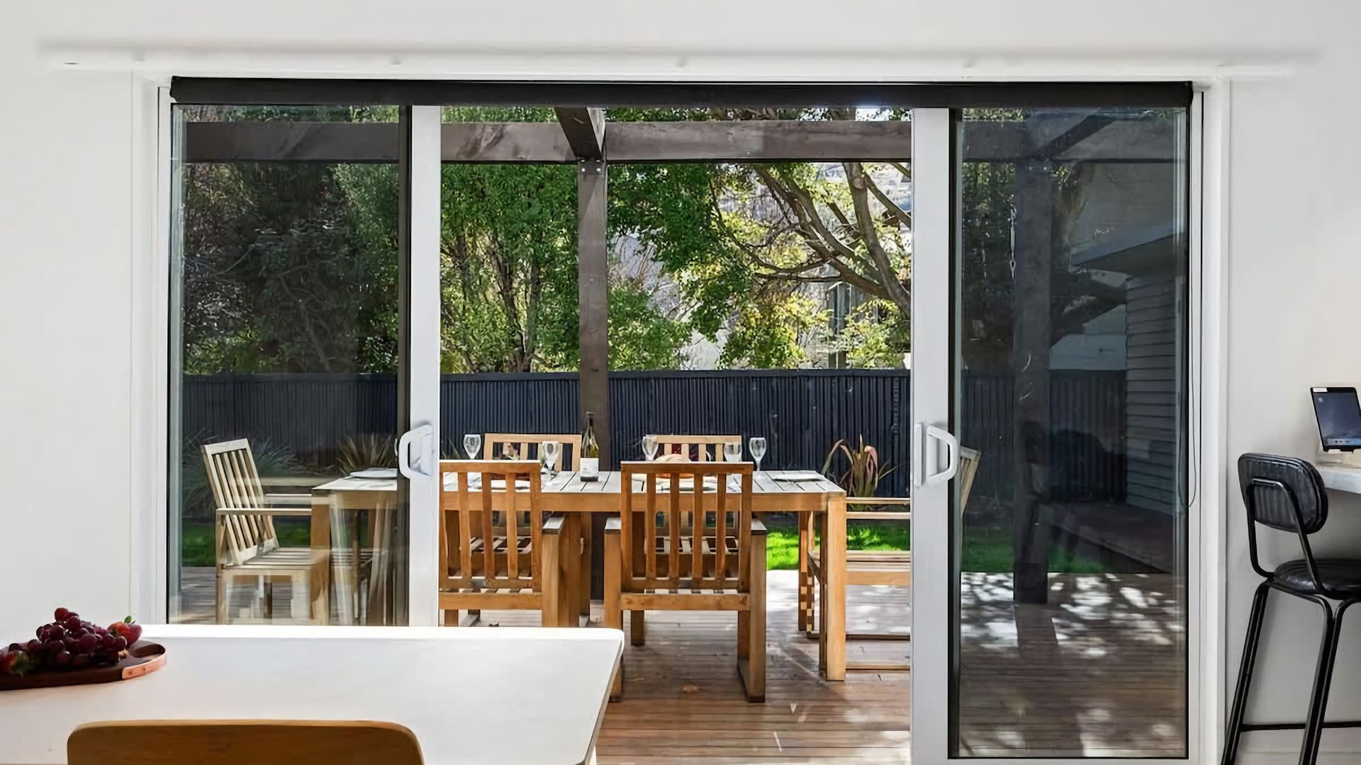 A dining area with a wooden table and chairs, a glass sliding door leading to an outdoor space with a wooden deck, and a laptop on a table to the right.