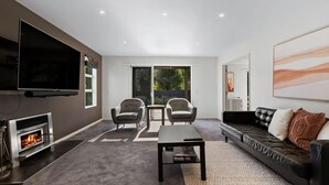 Een moderne woonkamer met een open haard, een flatscreen televisie, twee fauteuils, een bank en een groot raam met uitzicht op groen.