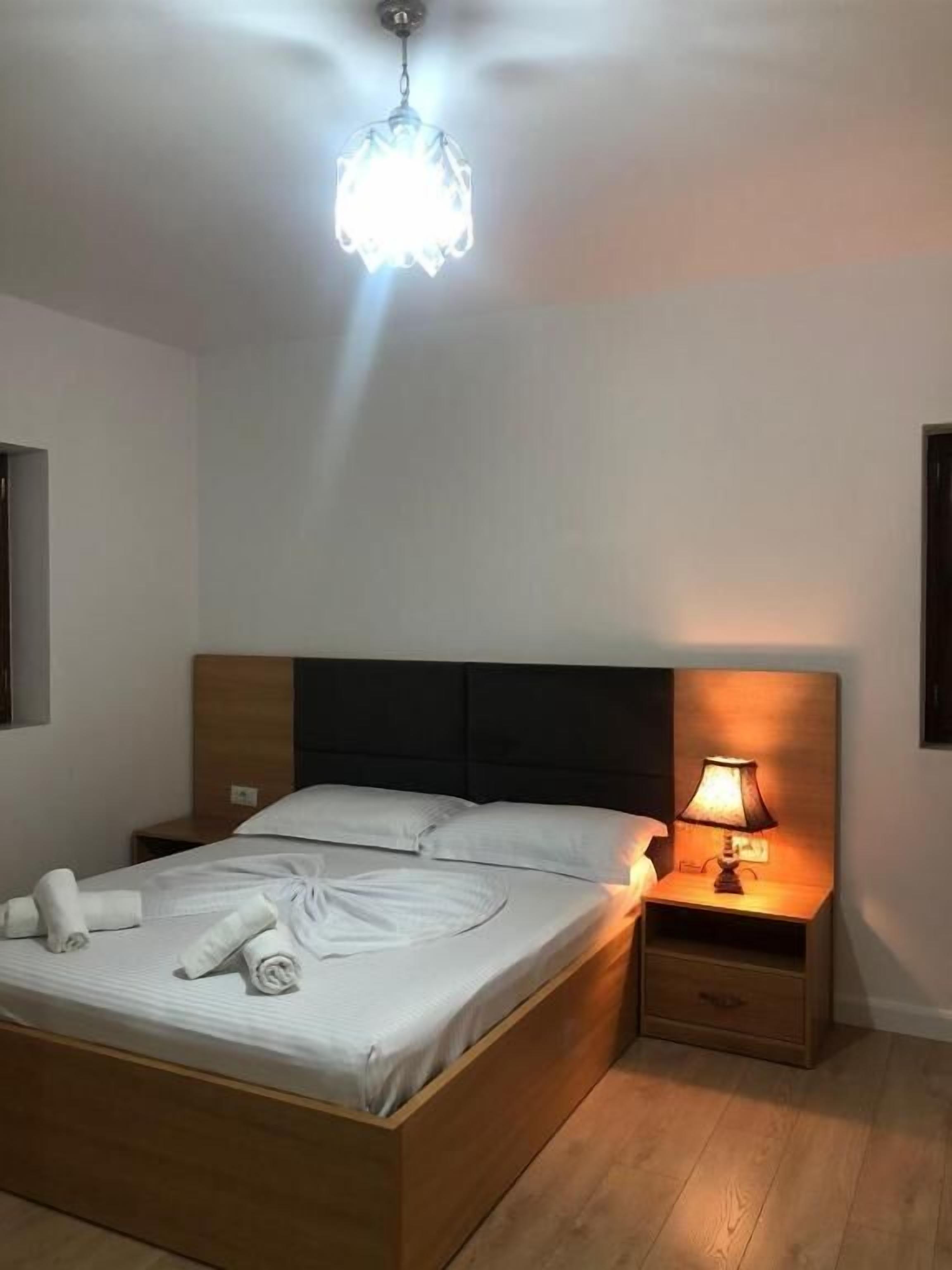 Een net opgemaakt bed met witte lakens en opgerolde handdoeken, een houten hoofdbord, een bedlamp en een hangende lamp.