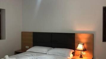 Een net opgemaakt bed met witte lakens en opgerolde handdoeken, een houten hoofdbord, een bedlamp en een hangende lamp.