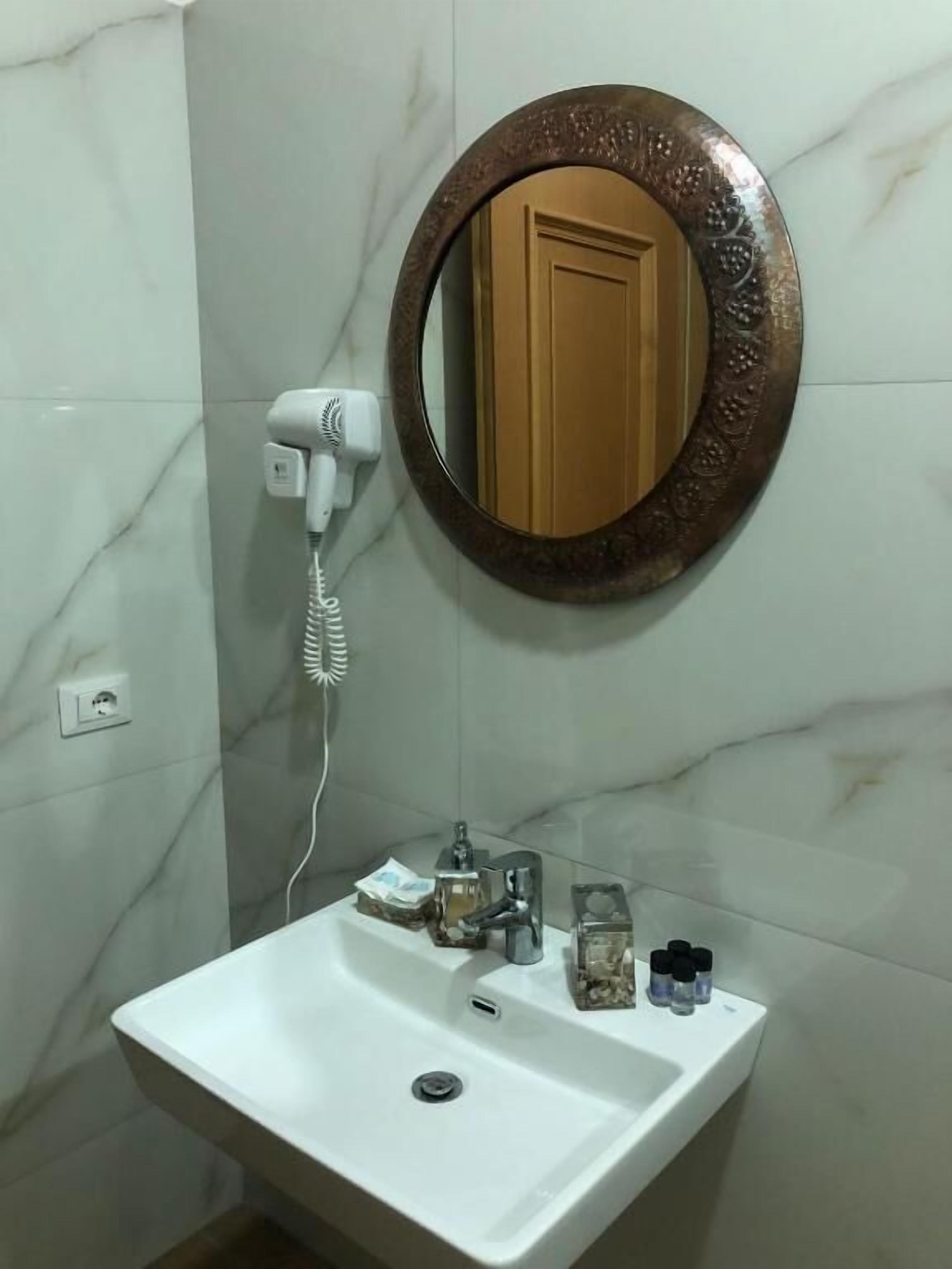 A bathroom with a marble wall, a round mirror with a decorative frame, a white sink, and a hairdryer on the wall.